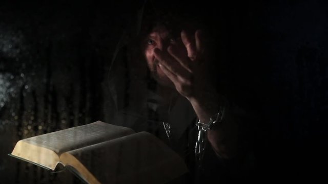 necromancer casts spells from thick ancient book by candlelight, behind transparent glass covered by water drops on a dark background