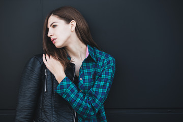 Portrait of brunette girl wearing leather jacket standing outdoors in the city against the black urban wall