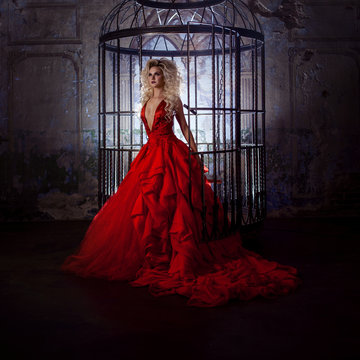 Fashion blonde in red dress with fluffy skirt near the birdcage, concept of liberation