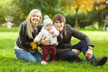 Naklejka na ściany i meble Happy parents with daughter sitting on grass in autumn park