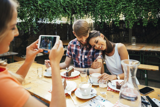 Woman taking cell phone picture of couple sitting outdoors with coffee and cake