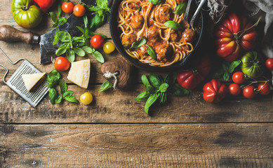 Italian pasta spaghetti with tomato sauce and meatballs in cast iron pan served with Parmesan cheese, fresh basil and colorful tomatoes over old rustic wooden background. Top view, copy space