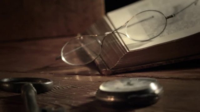 A pair of ancient glasses lying on an old book. Old wooden table with watch and a old key in the foreground. Camera panning and moving. Closeup.  Shallow depth of field.
