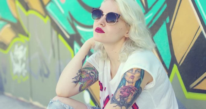 Trendy young woman with a pierced lip and vampire tattoo wearing stylish sunglasses sitting on a sidewalk leaning against a graffiti covered wall