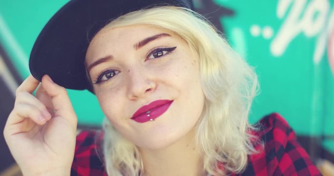 Gorgeous young blond woman with a pierced lip wearing a trendy baseball cap and crimson lipstick posing for the camera in front of colorful graffiti