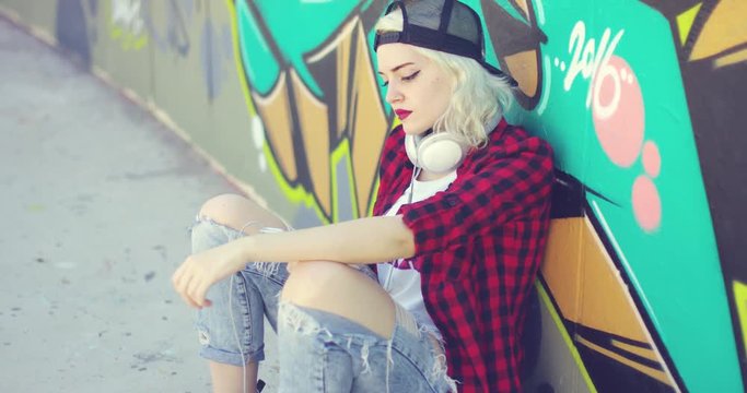 Trendy young urban woman wearing a baseball cap and torn jeans sitting waiting on her skateboard in front of a colorful graffiti covered wall
