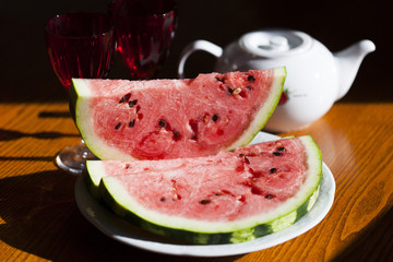 Куски арбуза на белой тарелке, на заднем плане - бокалы для вина из белого и красного стекла