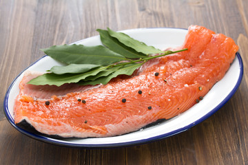 raw salmon with black pepper and bay leaf on white dish on brown wooden background