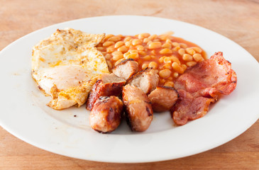 A fried English breakfast of sausges, bacon, fried and baked beams, on a white plate