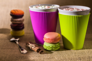 pink and green colored macaroons on each other and stick of cinnamon near spoon on foreground with two vivid colored cups of hot drink with marshmallows on burlap and stack macaroons on background
