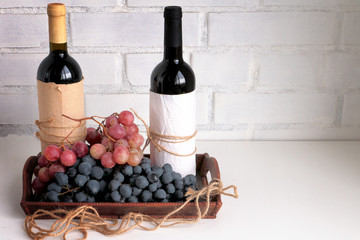 Bottles of dry red wine and bunch of dark red and purple grapes on white board against white brick...