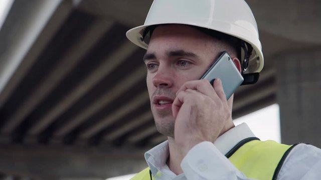 Handsome caucasian worker in white helmet and yellow vest talking on mobile phone while standing under overpass construction