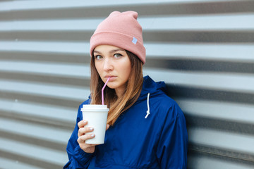 Stylish sporty brunette woman in trendy urban outwear posing with big white disposable cup straw cold rainy fall day against striped metal background. Vintage filter film saturated color.