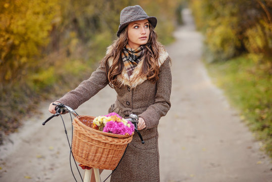 Beautiful girl in brown autumn coat and hat carrying ladies bicycle with basket full of flowers and autumn trees on the background.