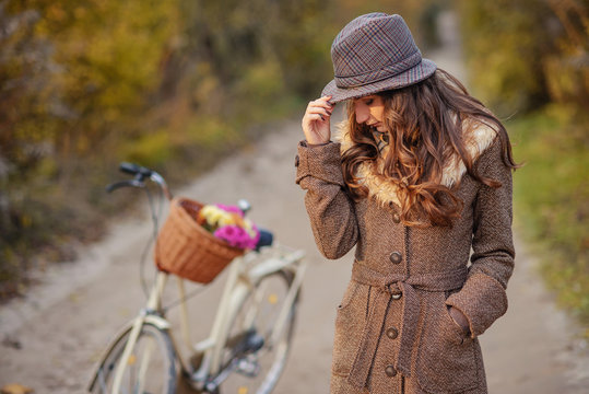 Beautiful girl in brown autumn coat and hat standing on the dirt road near the ladies bicycle and autumn trees on the background.