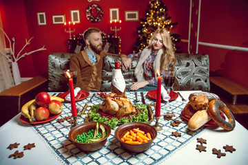 Beautiful couple in a decorated festive interior with a Christmas tree drinking wine. A romantic dinner for thanksgiving with fried chicken and candles