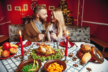 Beautiful couple in a decorated festive interior with a Christmas tree drinking wine. A romantic dinner for thanksgiving with fried chicken and candles