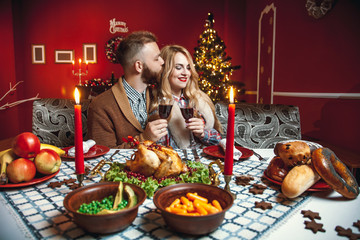 Beautiful couple kissing and holding glass of wine in a decorated festive interior with a Christmas tree. A romantic dinner for thanksgiving with fried chicken and candles