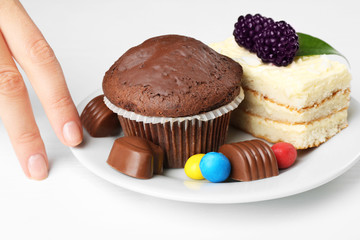 Female hand and plate with tasty sponge-cake, chocolate muffin and sweets on white table. Diet interruption concept