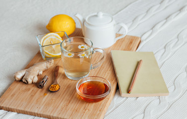 Obraz na płótnie Canvas Herbal tea with chamomile flowers, turmeric and honey on a wooden board. Treatment of hot drink with ginger. Treatment of folk remedies in bed. Leisure with a book.