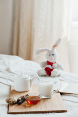 Hot milk in a glass cup and honey on a wooden board. Treatment of children a hot drink. Treatment of folk remedies in bed. Soft toy bunny for your child.