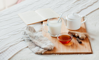 Hot milk in a glass cup and honey on a wooden board. Treatment of children a hot drink. Treatment of folk remedies in bed. Warm knitted socks. The book to read.