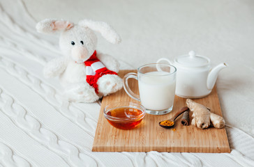 Obraz na płótnie Canvas Hot milk in a glass cup and honey on a wooden board. Treatment of children a hot drink. Treatment of folk remedies in bed. Soft toy bunny for your child.