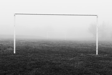Black and white image of soccer football goal posts in empty field pitch in winter fog