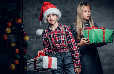 Attractive teenage boy on Santa's hat and cute blonde girl holds Christmas gift boxes.