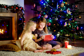 Happy little sisters opening magical Christmas gift by a fireplace