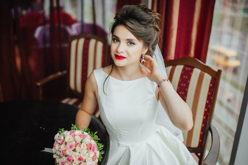 Naklejka premium Beautiful brunette Bride wedding Portrait. Red lips makeup. Wavy hair style. Bridal bouquet of flowers. Elegant lady sitting in red interior.