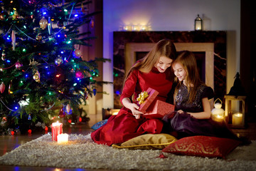 Young mother and her daughter unwrapping Christmas gifts by a fireplace in a cozy dark living room on Christmas
