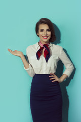 Beautiful dark haired young business woman dressed in a dark blue suit with a red, blue scarf standing and showing to her right side with hand, isolated on blue background. Stewardess