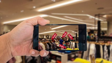 Fototapeta na wymiar Augmented reality application for retail business concept. Hand holding smart phone with A/R application on screen to scanning sale alert in men wear shop in department store. 