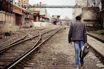 Young man with guitar case is going away among industrial ruins
