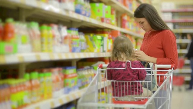 Mother and child walks along wholesale shelves and taking goods in shop trolley, beautiful young wife shopping in a supermarket