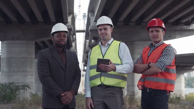 4K shot of front view of three architects one african and two caucasians in protective helmets hard hats looking at camera.