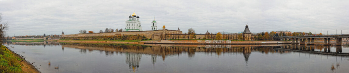 Fototapeta na wymiar Panoramic view of Pskov Kremlin. Trinity Cathedral in old fortress. Autumn cityscape wide panorama. Translation of the inscription on the bank: Russia starts here.