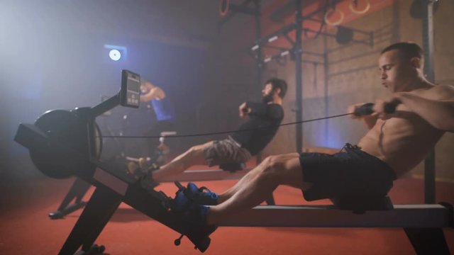 Three athletes doing rowing and air cycling crossfit exercises at the gym.