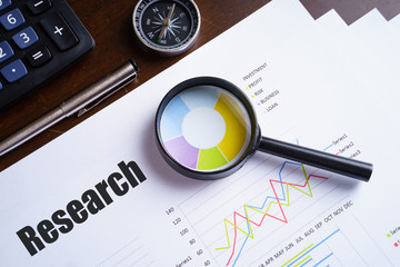Magnifying glass on "Research" text on chart, dice, spectacles, pen, laptop calculator on wooden table - business, banking, finance and investment concept