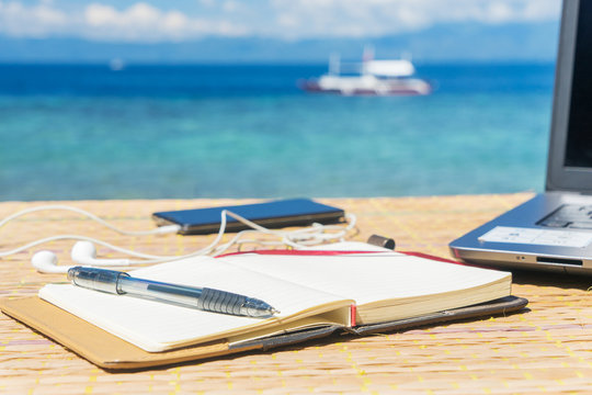 Opened empty notepad is on the table with phone and headphones at the tropical sea background