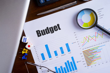 "Budget" text on paper sheet with magnifying glass on chart, dice, spectacles, pen, laptop and blue and yellow push pin on wooden table - business, banking, finance and investment concept