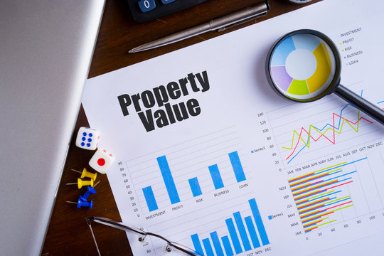 "Property Value" text on paper sheet with magnifying glass on chart, dice, spectacles, pen, laptop and blue and yellow push pin on wooden table - business, banking, finance and investment concept