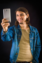 Man in denim jacket makes selfie on a smartphone. With long hair and a beard. On a black background.