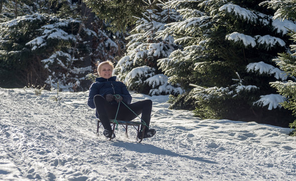A smiling middle-aged blonde goes from a hill on a sled on a background of snow-covered forest. Selective focus.