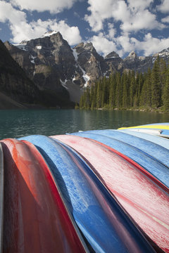 Row Of Canoes On Their Sides With A Lake Mountains Clouds And Blue Sky; Alberta, Canada