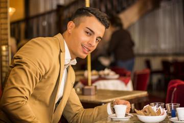 Portrait of smiling handsome man in jacket sitting at table with cup of drink looking at camera