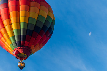 This is a photo of a beautiful hot air balloon slowly sailing through a calm blue sky showing the moon in the background