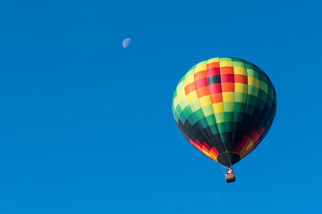 This is a photo of a beautiful hot air balloon slowly sailing through a calm blue sky with a moon in the background.