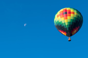 This is a photo of a beautiful hot air balloon slowly sailing through a calm blue sky with the moon in the background.